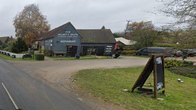 Single-story pub painted grey. The words "The Rusty Gun" and "Restaurant" are visible in white lettering. A pub A-frame is visible in the foreground, alongside a driveway edged in grass. A road passes the pub to the left side.