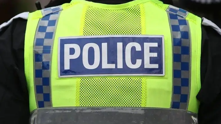A police is standing with their back towards the camera. They are wearing a black jacket and a yellow hi-vis that says POLICE on the back.
