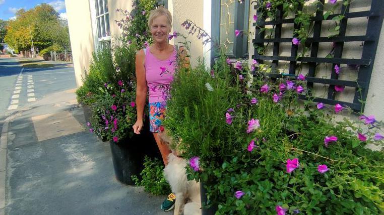 A woman with blonde hair and a full fringe tied back is wearing a bright pink vest and blue and pink shorts. She is smiling at the camera and standing next to a black and silver front door. Next to the front door are several purple flowers and greenery that she has planted. You can see the pathway in front of her and a beige-coloured dog is sat next to her, looking up at her.