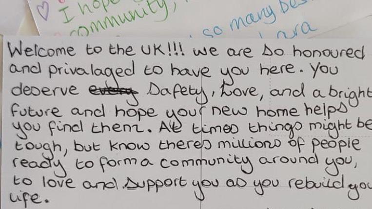 A note with a handwritten message that reads Welcome to the UK! We are so honoured to have you here. You deserve safety, love, and a bright future and hope your new home helps you find them. At times things might be tough, but know there's millions of people ready to form a community around you, to love and support you as you rebuild your life.