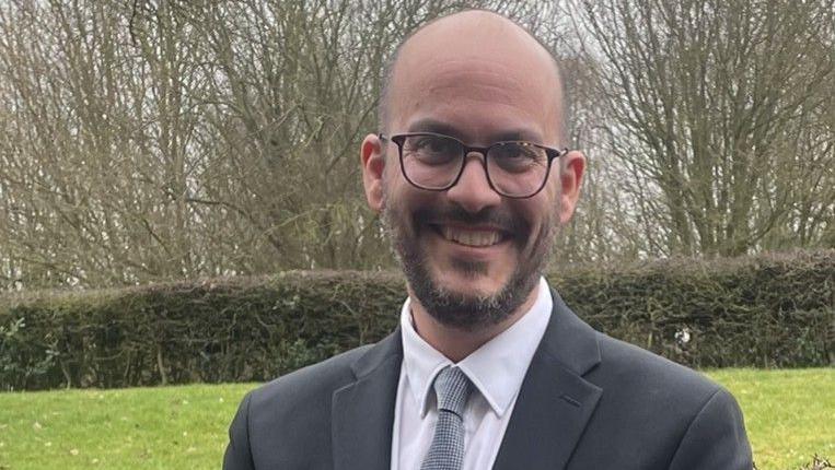 Maughan Johnson, smiling at the camera, standing outside, wearing a grey suit, with a white shirt, grey tie, he has on glasses, has a dark beard and a bald head. Grass, bushes and trees are behind him.