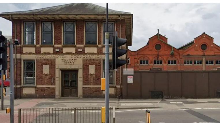 A red bricked two-storey building with the words Northampton Corporation Transport Offices above the door. It stands on the corner of a road in front of a pedestrian crossing.