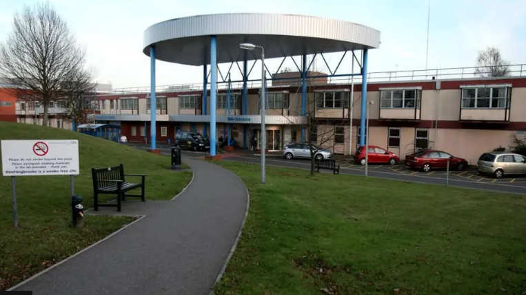 View of two-storey Hinchingbrooke Hospital, showing lawn, a pathway, a line of parked cars and a covered area over the main entrance 