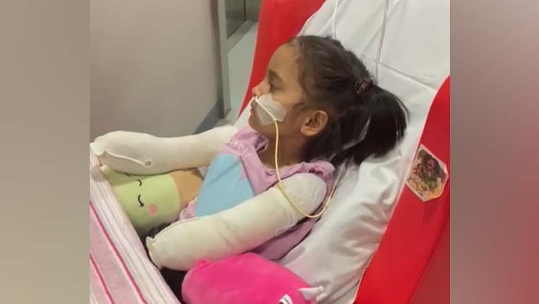 Saffanah is in a hospital bed. She has a tube connected to her nose and bandages all the way up her arms.