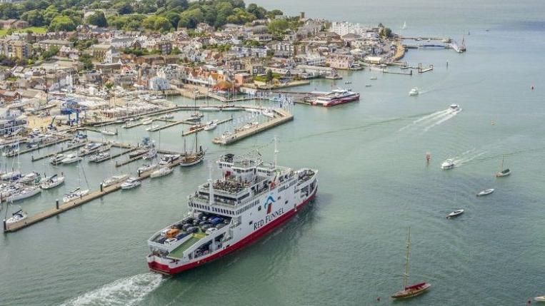 A stock image of a Red Funnel ferry in the sea, near a port with houses and other boats around the ferry and by the port too. 
