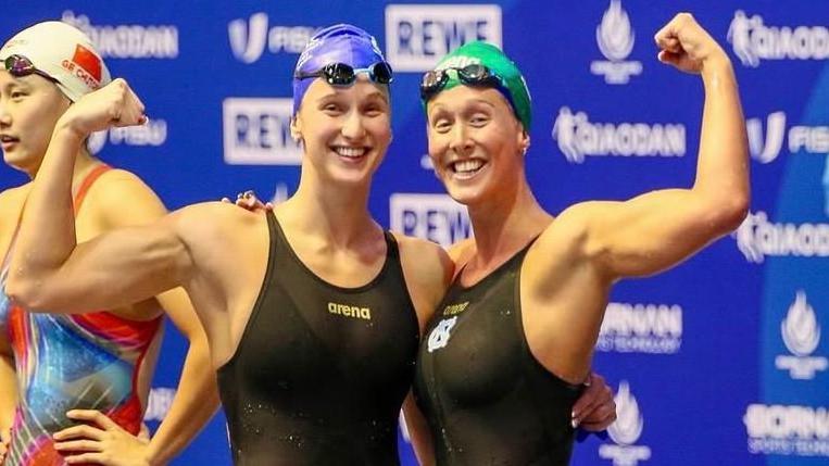 Olivia and Georgia Nel in black swimming costumes after a race, with their arms held aloft.