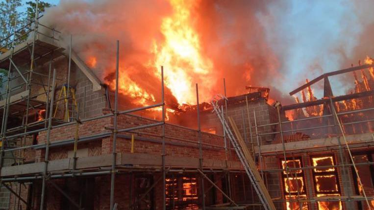 Flames and smoke inside and from roof of house partially built - it is surrounded by scaffolding and is built with large grey blocks/bricks.