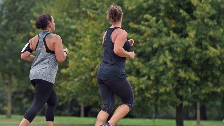 Two women jog through a leafy green park. Both wear vest tops and leggings. One of the women, on the left, also wears a running armband to hold her phone.