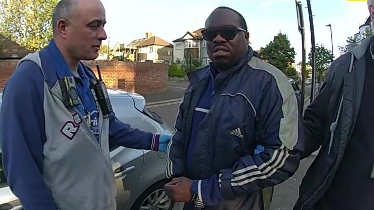 Bodycam footage showing Simon Levy in a navy blue Adidas jacket and sunglasses being detained by police officers on a suburban street. An officer on the left wearing a blue shirt and grey jacket, fitted with a body-worn camera and radio, holds the man's arm.