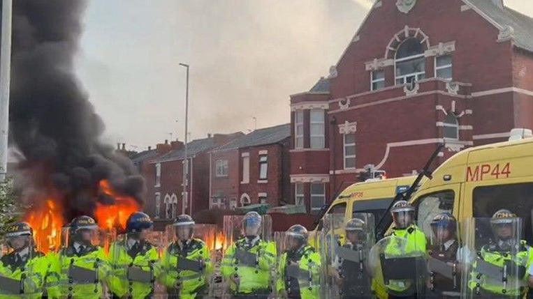 A line of police officers in riot gear stands in front of the Southport mosque where rioting broke out in July as cars and bins are blazing.