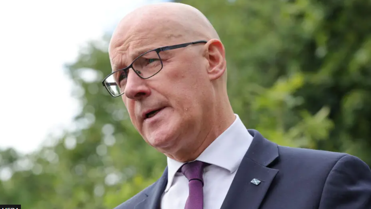 Prìomh-mhinistear na h-Alba, John Swinney. Tha e maol agus tha speuclairean air. Tha seacaid ghorm agus tàidh purpaidh air. 