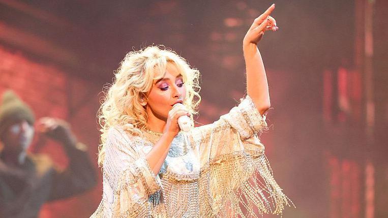 Sabrina Carpenter performs in a white kaftan-style top with long gold tassles dangling from each sleeve. She sings into her signature jewel-encrusted microphone, her free hand pointing upwards. In the background a dancer in overalls waves their arms.