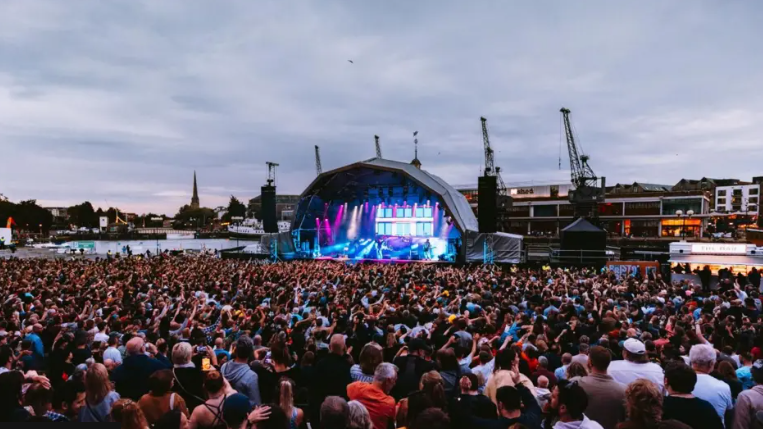 A crowd of people watch a musical act on a lit up stage on Bristol harbourside with the dock's cranes in the background