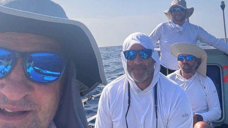 Four rowers in hats and white tops and wearing sunglasses in a boat with sea behind.