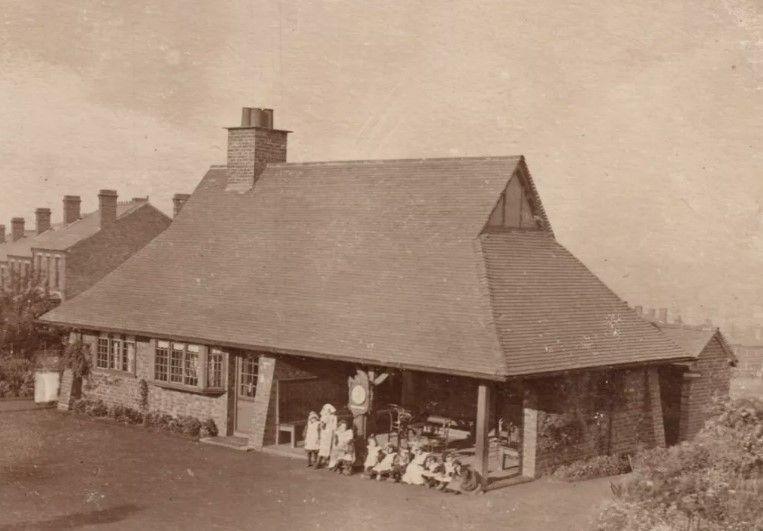 A black and white photo showing a public building on Bury Hill Park with a row of young children sitting on a step along the front.  