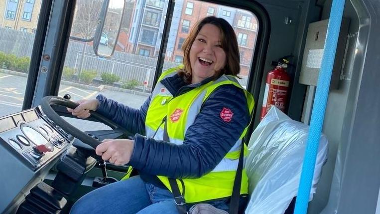 A woman in a yellow high-vis jacket on top of a blue winter coat, both with The Salvation Army branding on them, smiling at the camera. She is in the driver's seat of a bus.