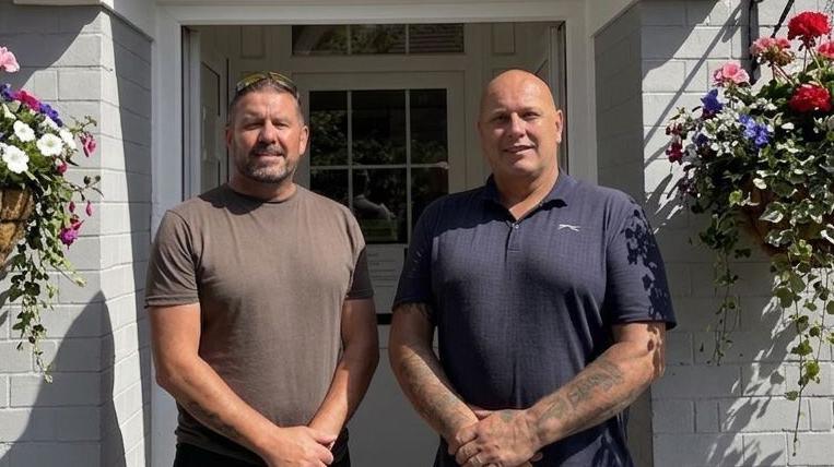 Two men smiling at the camera outside a white building. The one on the left has a brown t-shirt on and short brown hair and has his arms in front of him with his hands clasped together. The one on the right is bald with a blue t-shirt on, with his arms in front of him and his hands clasped together.