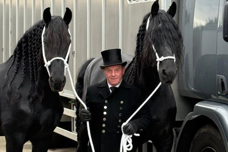 Modris Kesans with two black horses either side of him. He has a black jacket on with silver buttons and a black top hat. He holds the horses with white rope.
