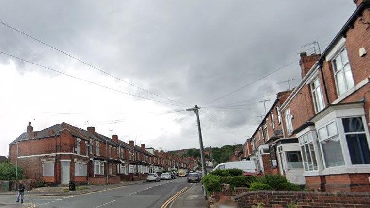 A road with detached houses lining each side and- several vehicles parked on the street.