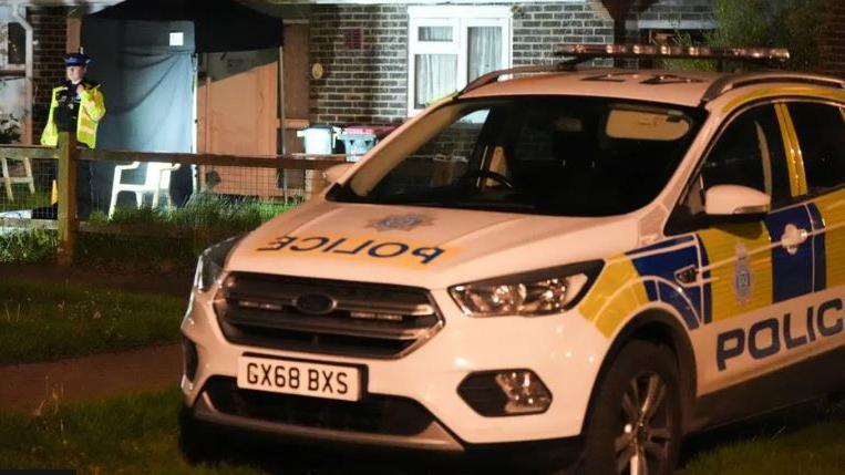 A police car parked outside a bungalow with a police officer stood outside the front door in low light