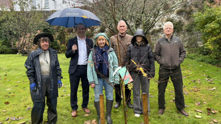 A group of people dressed in raincoats standing in a garden. They are standing behind a small tree. They are smiling at the camera.
