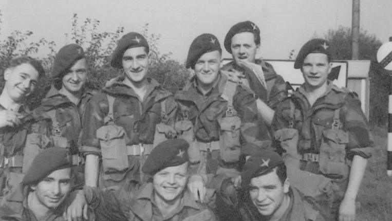 A black and white photo of WW2 troops standing side by side, some with their arms around each other, in full military uniform.