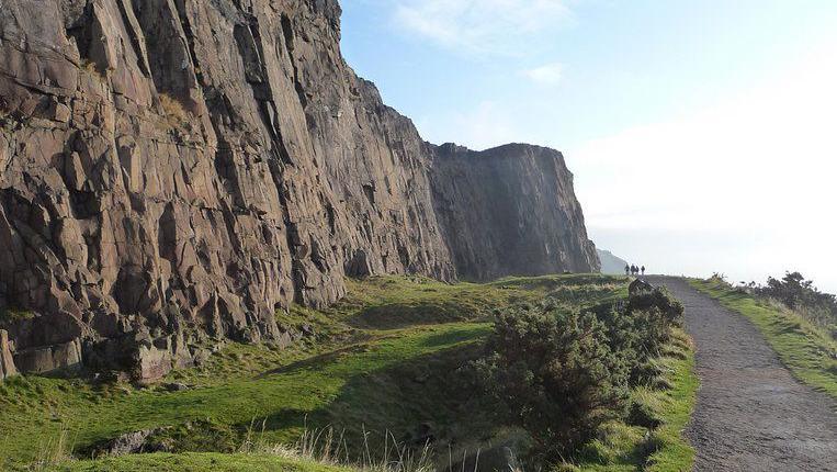 The steep rocky crags rise above the road. A group of people are walking in the distance along the Radical Road.