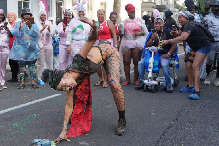 A woman is bent over as she dances in the street. She wears fishnet tights and a black top and head scarf. People look on and smile, some with their cameras pointing at the woman. 