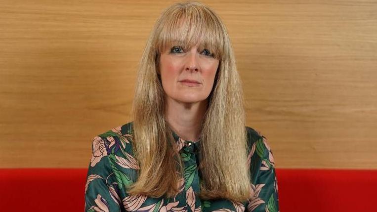 This image shows the Children and Young People's Commissioner for Scotland Nicola Killean. She has a serious expression. She has blonde hair with a fringe and is wearing a green floral top.