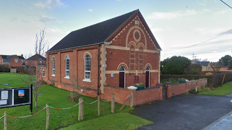 Ealand Methodist Church. The red brick building has three arched windows down the side and two doors at the front. It is set back slightly from the road and a church noticeboard can be seen to the left of the picture. 