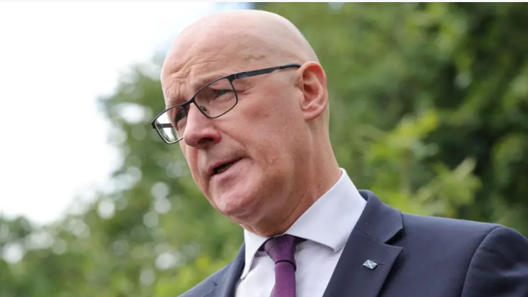 Prìomh-mhinistear na h-Alba, John Swinney. Tha e maol agus tha speuclairean air. Tha seacaid ghorm agus tàidh purpaidh air. 