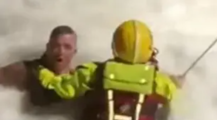 A man in the sea is held by a firefighter wearing a yellow helmet and rescue gear with rope attached going off to the right of the photo.