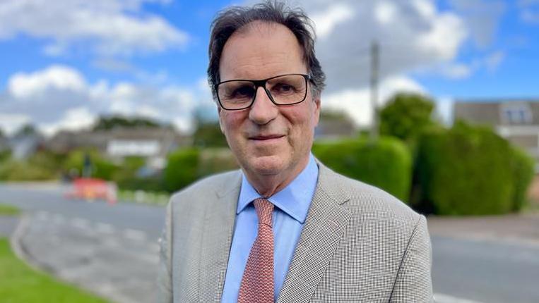 Henry Cator standing in Mill Lane in Attleborough, which was flooded during Storm Babet. He is wearing glasses and he is in a checked, light grey jacket. He is wearing a blue shirt with a patterned tie. The background is out of focus.