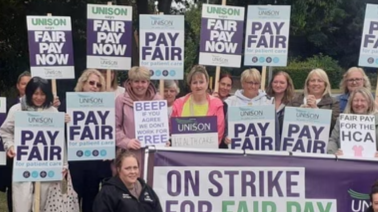 Staff with "pay fair" placards and a banner standing on grass outside the hospital