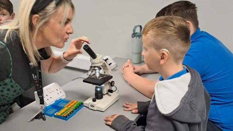 A woman with shoulder-length blonde hair and a headband and black top is using a microscope and looking at a young boy, who has short blonde hair, a grey hoody and dark trousers. He is stood next to a boy with short brown hair, blue top and dark trousers.