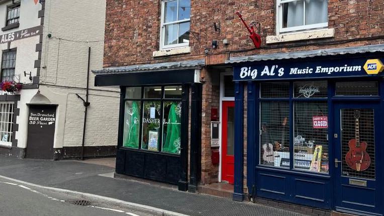 Picture of the 'Big Al's Music Emporium' shop front - it's painted navy blue with the name written in white lettering. In the front window there's a large burgundy guitar hung up, a ukulele, and a Casio keyboard.