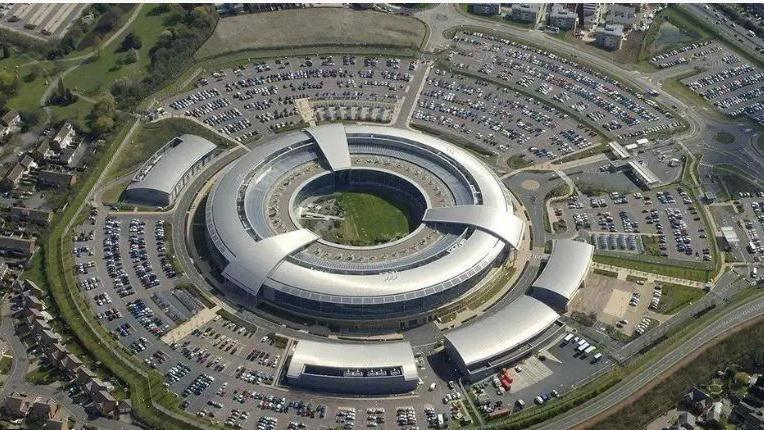 An aerial view of GCHQ headquarters, a circular, silver metal coloured building with several small units nearby, surrounded by car parks, a public road and a housing estate.