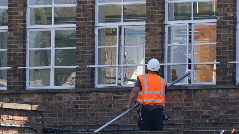 A man has his back turned to the camera, wearing a white hard hat and orange high-vis vest. He is holding a long metal pole and is standing at the foot of a school building. He is possibly standing on a flat roof.