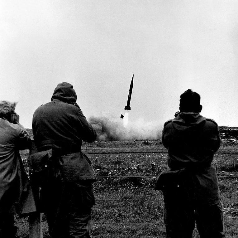 Three men look through binoculars from behind a wire fence as a missile takes off. There are flames emerging from the rocket and a plume of smoke.