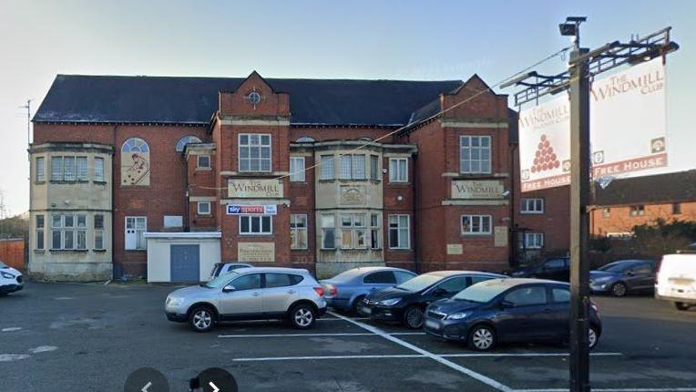 Two storey brick-built social club with bay windows on both floors to the left and right. There are white or cream surrounds to the windows,. There are cars parked in painted spaces in front of the building. A pub sign with red letters on a cream background is visible to the right.