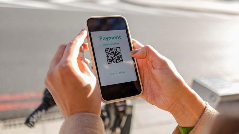 A pair of hands are holding a mobile phone in a car park. There is a QR code on the screen of the phone. 