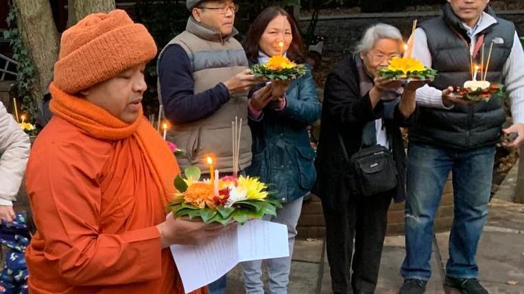 The monk is dressed in orange drapes and wears an orange hat while holding a kratong and performs prayers. Four worshippers on the right of him join in. 