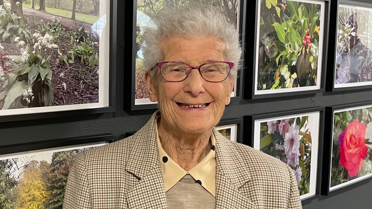 Betty, stood smiling in the exhibition. Behind her is a wall of framed photographs - we can see close-up shots of plants and pink flowers.