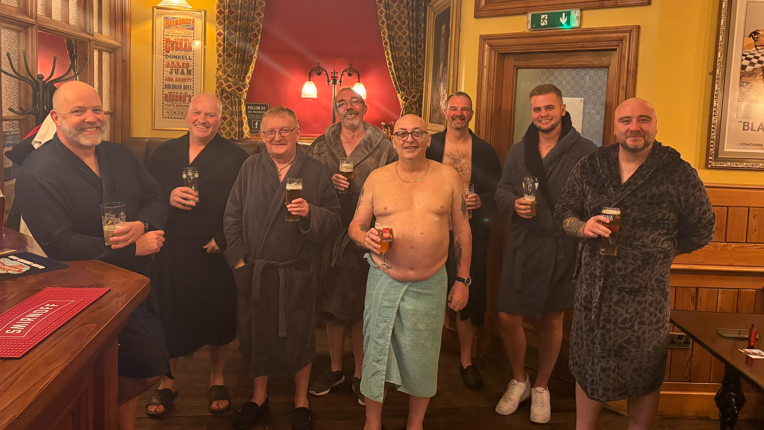 A group of men in dressing gowns and towels in a pub, each holding a pint.