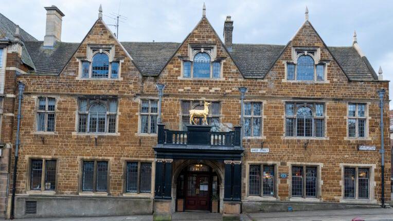 Historic stone building with ornate architecture, featuring multiple pointed gables, large mullioned windows, and a central entrance with a black balcony topped by a golden hind statue. The building has signs reading 'The Hind Hotel' and is situated along a paved street with two red bollards in the foreground.