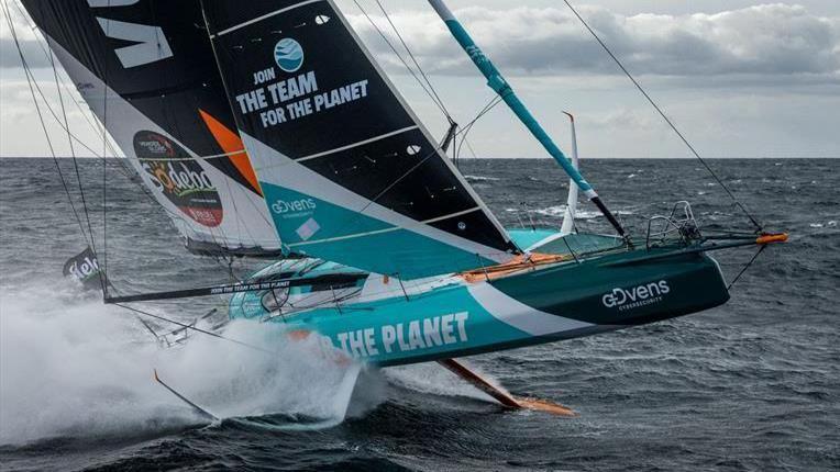 A racing yacht breaching the water with foils either side of the hull and a long centreboard underneath with blue white and black sails.