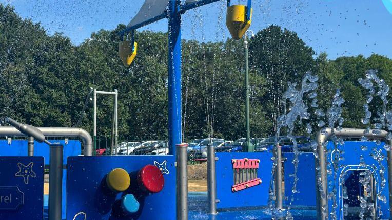A splash park which has a blue floor and there are blue plastic panels propped up vertically by silver grey poles. There are jets spraying water into the air.