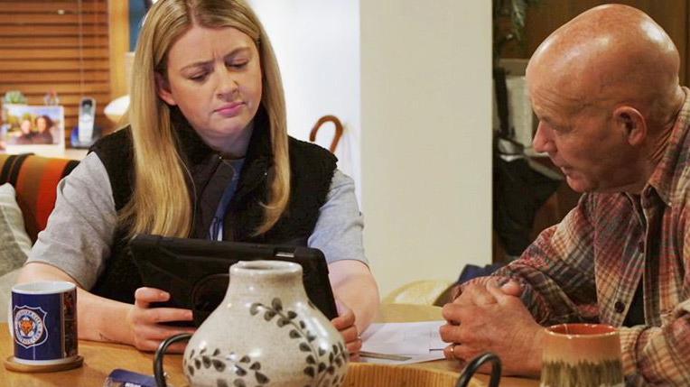 Two actors, one female and one male, sat at a table looking at a tablet with a Leicester City mug visible on the table.