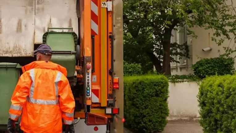 A bin crew member wearing an orange jacket and a black cap- standing in front of an orange lorry emptying green bins