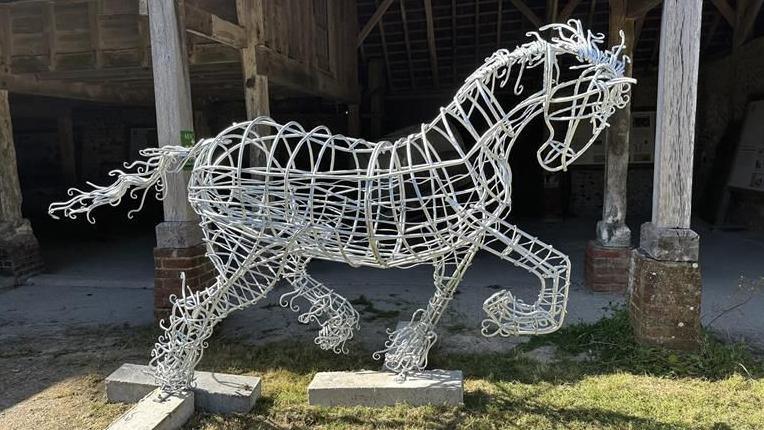 A sculpture of a running horse made out of steel. The metal is a light silver colour and the sculpture is not solid, with the main body made out of mostly circular pieces of metal with rods attaching them together. Three of its four hooves are bent and in the air, making it appear as if it is trotting or cantering. The horse's tail has been made with curvy pieces of metal, creating a flowy effect. The sculpture is on a patch of grass in front of what appears to be a barn.